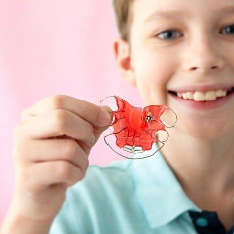 Cheerful child with metal braces smiling and holding a red orthodontic retainer against a pink background