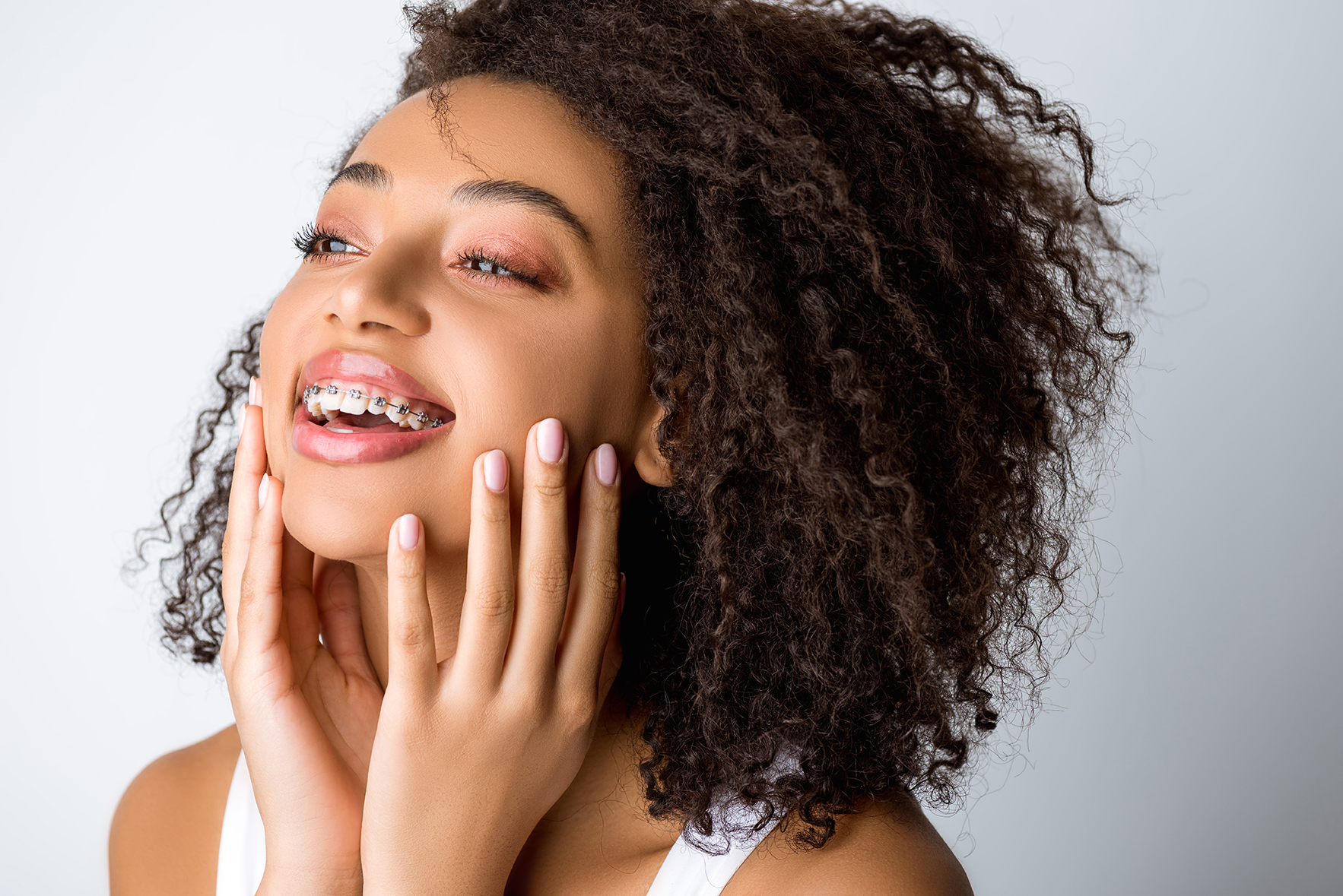 Young woman with curly hair and metal braces laughing while touching her face against a grey background